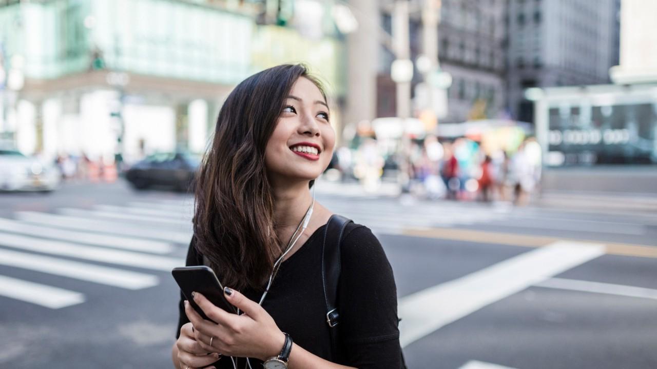 Smiling woman crossing the street in a big city, with a smartphone in her hand and ear plugs.